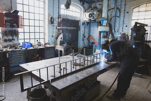 A blacksmith wears safety gear and is welding a metal construction in a metalsmith's workshop.