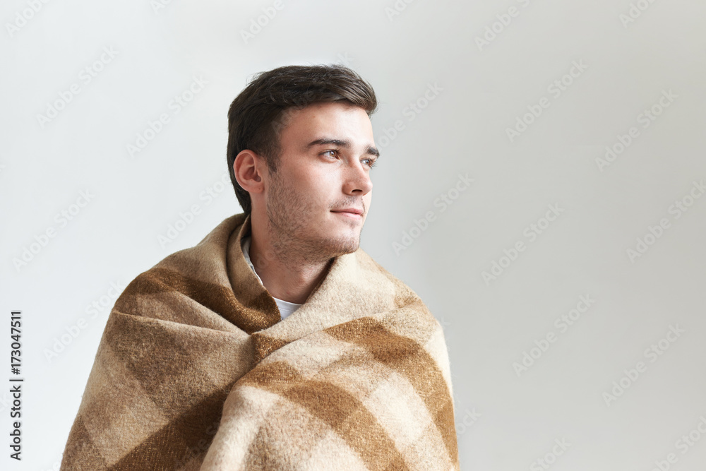 Picture of handsome young male with stubble spending winter morning indoors, wrapping in wool plaid, feeling positive about new day, looking sideways with happy dreamy expression on his face Picture of handsome young male with stubble spending winter morning indoors, wrapping in wool plaid, feeling positive about new day, looking sideways with happy dreamy expression on his face