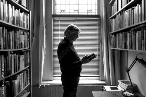 Senior man reading a book standing between bookshelfs photo