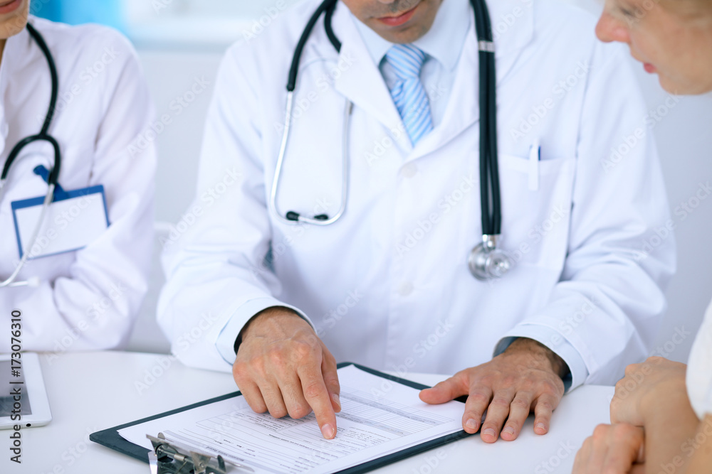 Doctor and patient talking while sitting at the desk in office. Physician pointing into medical history form. Medicine and health care concept Doctor and patient talking while sitting at the desk in office. Physician pointing into medical history form. Medicine and health care concept