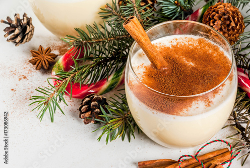 Traditional winter and Christmas drink eggnog with grated nutmeg, anise stars and cinnamon. On white marble table, with spices, christmas tree, pine cones, candy cane, copy space