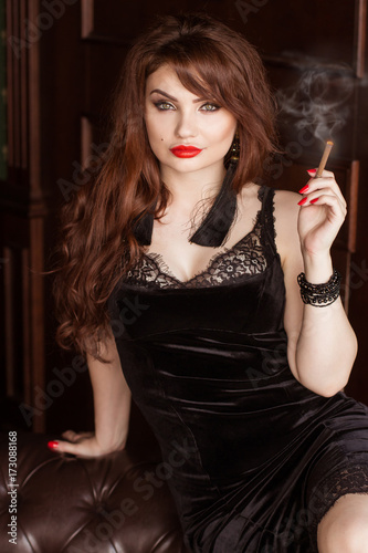 A beautiful sexy woman in a black dress smokes a cigarette and drinks whiskey. Sofa chesterfield. Mistress, Spy Girl