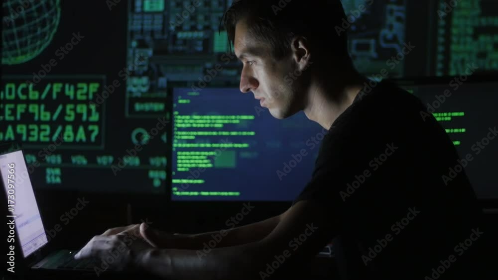 portrait of a young programmer working at a computer in the data center filled with display screens