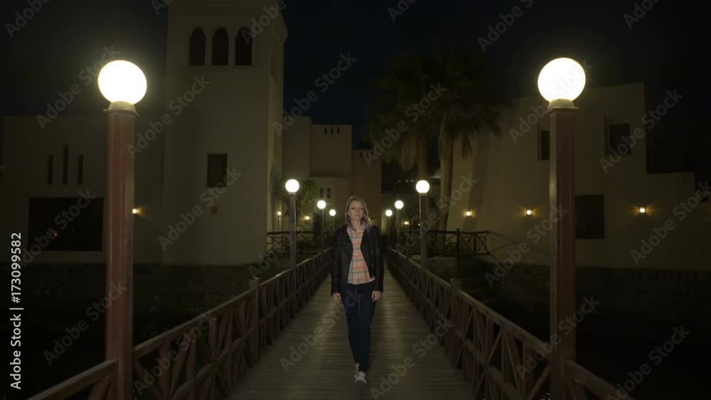 Blonde young woman in leather jacket walks alone on wooden pedestrian bridge between lamp post at night or evening. Sad beautiful girl in sorrow, dreams, loves, misses, thinks about meaning of life Blonde young woman in leather jacket walks alone on wooden pedestrian bridge between lamp post at night or evening. Sad beautiful girl in sorrow, dreams, loves, misses, thinks about meaning of life
