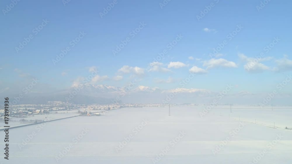 AERIAL: Flying above snowy fields and suburban town, high rocky mountains in background. Snow covered houses in wintry countryside city on misty morning. Snowy scenery of quiet suburbs and snowy Alps AERIAL: Flying above snowy fields and suburban town, high rocky mountains in background. Snow covered houses in wintry countryside city on misty morning. Snowy scenery of quiet suburbs and snowy Alps