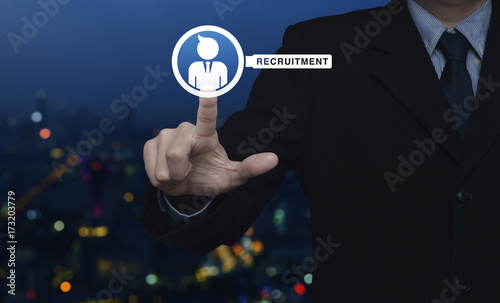 Hand pressing businessman with magnifying glass icon over blur colorful night light city tower, Business recruitment concept