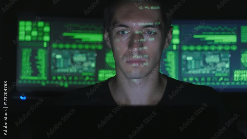 Hacker programmer working on a computer while green code characters reflect on his face in a dark office room. Hacker programmer working on a computer while green code characters reflect on his face in a dark office room.