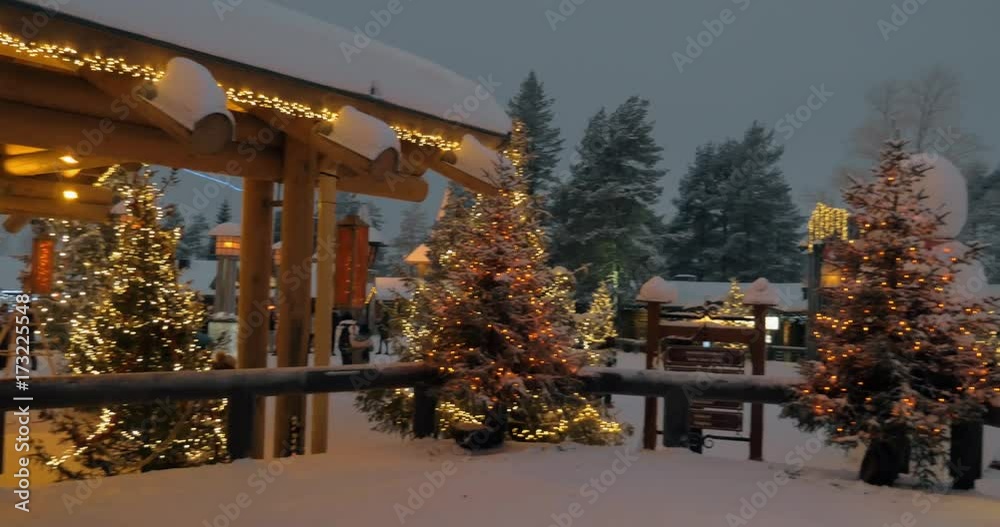 ROVANIEMI, FINLAND - JANUARY 06, 2017: Panning evening shot of Santa Village in Lapland. People walking among pines and wooden houses including Santa Office decorated with Christmas lights