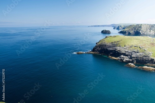 An aerial view from clifftops looking out to the horizon of a deep, blue, calm ocean at Tintagel in Cornwall, UK which is the seat of the legendary English King Arthur and shows cliffs.