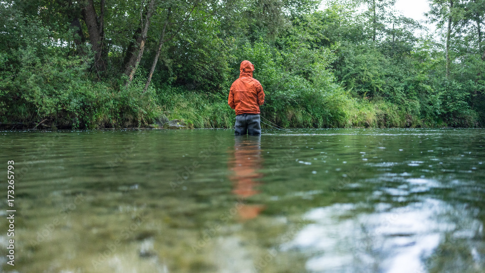 Angler mit Wathose und Watjacke im Wasser beim Angeln mit Fliegenrute bei Regen im klaren Fluss stehend Angler mit Wathose und Watjacke im Wasser beim Angeln mit Fliegenrute bei Regen im klaren Fluss stehend