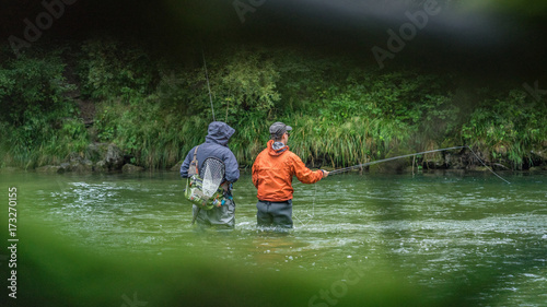 Angler mit Wathose und Watjacke im Wasser beim Angeln mit Fliegenrute bei Regen im klaren Fluss stehend und werfend