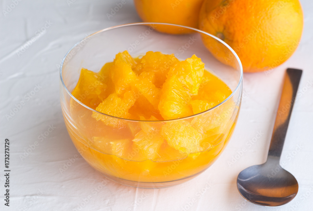 Fresh orange fruit slices with juice in glass dessert bowl on white rustic surface background and two whole oranges defocused Fresh orange fruit slices with juice in glass dessert bowl on white rustic surface background and two whole oranges defocused