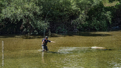 Angler mit Wathose im Wasser beim Angeln mit Fliegenrute bei Sonne im klaren Fluss stehend und werfend