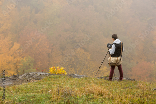 nature, work, hobby concept. young man dressed in warm clothes, burgundy pants, grey sweatshirt and black vest, standing next to tripod with camera and taking pictures of autumn forest