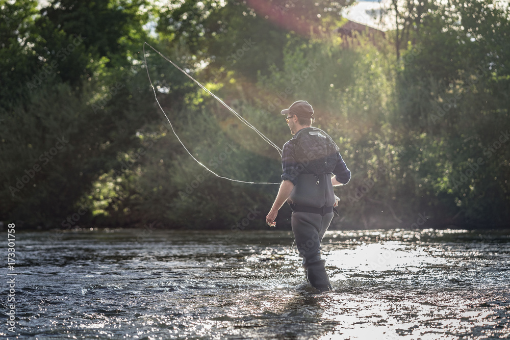 Angler mit Wathose im Wasser beim Angeln mit Fliegenrute bei Sonne im klaren Fluss watend Angler mit Wathose im Wasser beim Angeln mit Fliegenrute bei Sonne im klaren Fluss watend