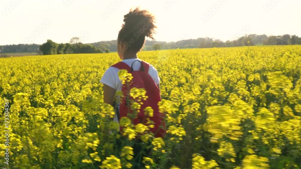 4K video clip of beautiful happy mixed race African American girl teenager female young woman hiking with red backpack in field of rape seed yellow flowers