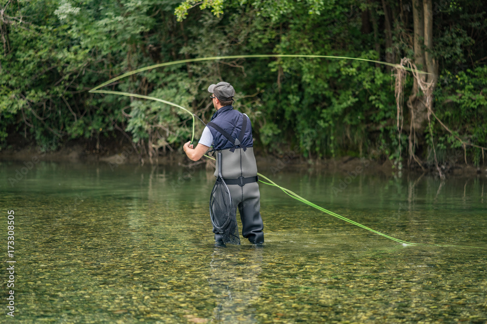 Angler mit Wathose und Fliegenrute im Wasser beim Angeln bei Sonne im klaren Fluss stehend und werfend Angler mit Wathose und Fliegenrute im Wasser beim Angeln bei Sonne im klaren Fluss stehend und werfend
