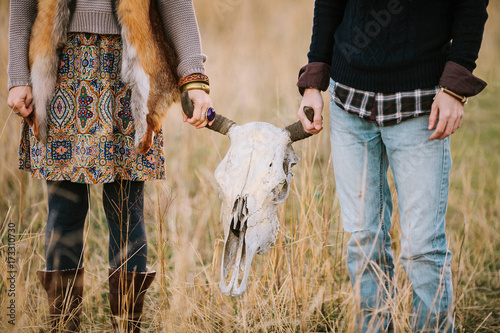 fall, fashion, youth concept. young man and woman dressed in boho style, she has ornamental skirt and vest made of fox skin, he is in light jeans and plaid shirt, they are holding big skull of bull