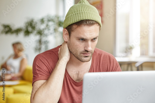 Serious concentrated young programmer in trendy hat doing reomote work, pondering and looking at laptop screen with thoughtful expression. Male copywriter working on publication for marketing project photo