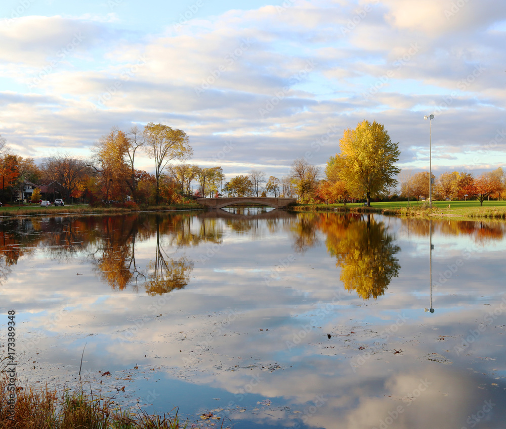Fototapeta premium Autumn nature background. Beautiful fall landscape in the city park. Colored trees and cloudy sky reflected in the water during sunset. Midwest USA, Wisconsin.