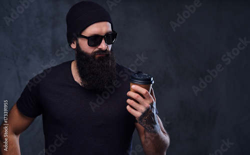 A man in a hat and sunglasses holds take away coffee cup.