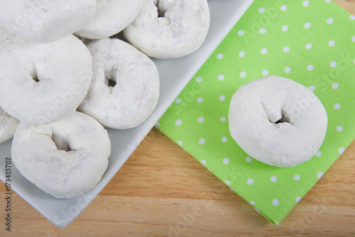 cropped angle view of a square plate with round powdered donuts on a light wood table, one donut isolated on green cloth napkin with white polka dots