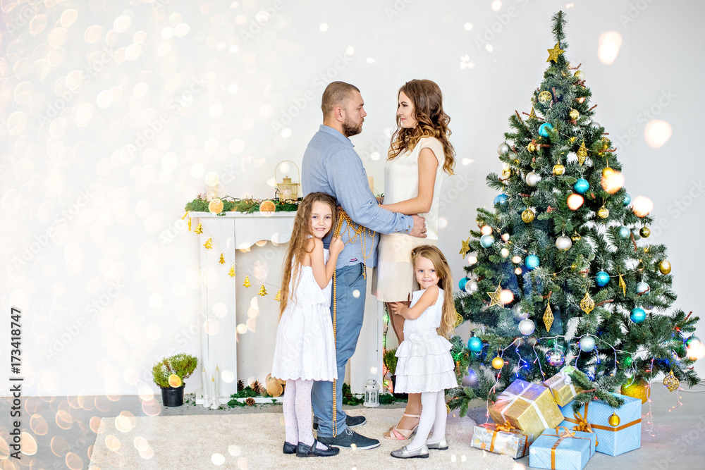 Happy family, parents hugging with their daughters by the white fireplace with an elegant Christmas tree in garlands of lights. A young father and a beautiful mother are holding hands, little sisters Happy family, parents hugging with their daughters by the white fireplace with an elegant Christmas tree in garlands of lights. A young father and a beautiful mother are holding hands, little sisters