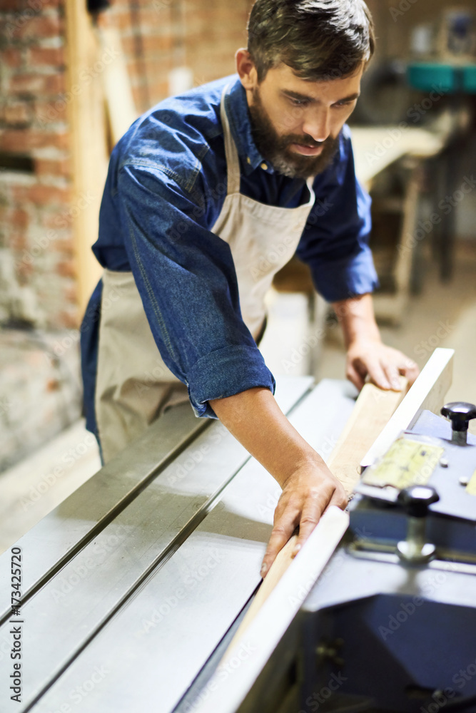 Portrait of bearded carpenter carefully placing piece of wood on cutting machine in modern workshop Portrait of bearded carpenter carefully placing piece of wood on cutting machine in modern workshop