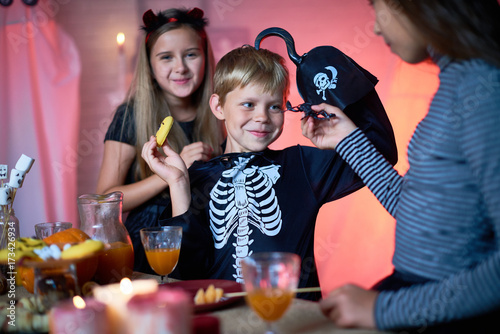 Girl showing toy scorpion to smiling cute boy in skeleton costume with hook instead of hand eating cookie at Halloween party, cheerful pretty girl behind him looking at camera