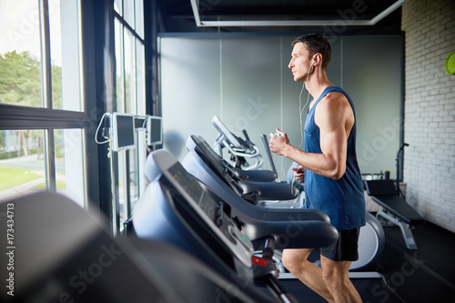Profile view of concentrated fit man listening to music in headphones while running on treadmill in modern gym with panoramic windows, portrait shot