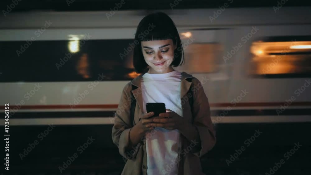 Beautiful young woman in trendy coat stands on platform in evening, smartphone lights up her face, she texts on messaging application, smiles and giggles, train passes by