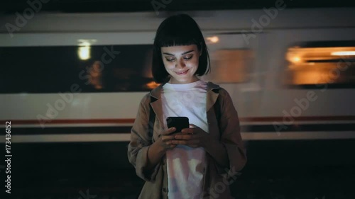 Beautiful young woman in trendy coat stands on platform in evening, smartphone lights up her face, she texts on messaging application, smiles and giggles, train passes by