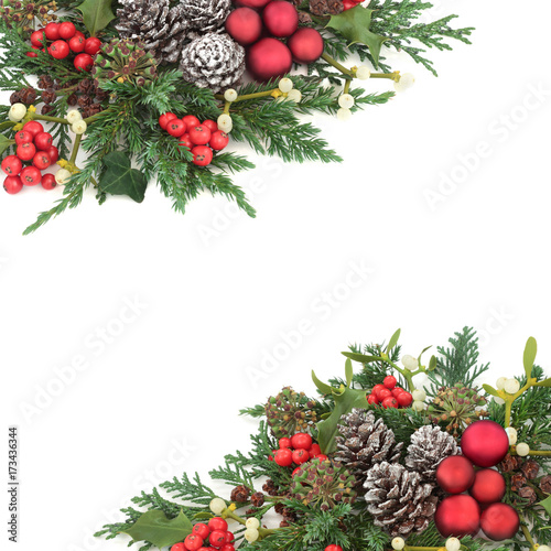 Christmas background border with red bauble decorations, holly, ivy, mistletoe, cedar and juniper leaf sprigs and pine cones over white. 