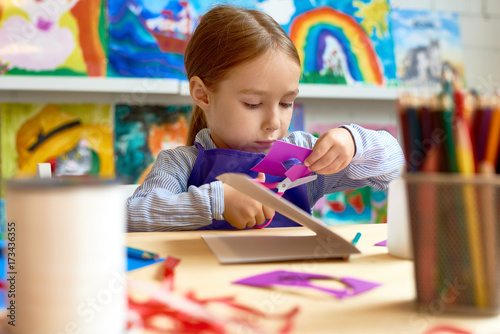 Portrait of cute little girl carefully cutting out shapes while making collage during art class in pre-school