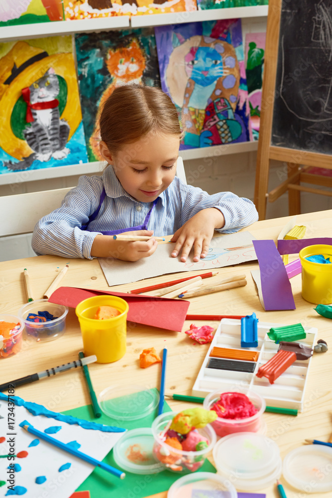 Fototapeta premium Portrait of adorable little girl writing wishes on handmade gift card sitting at messy table with supplies during art and craft class of pre-school