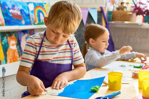 Portrait of cute little boy wearing apron making plasticine picture in art and craft class of pre-school