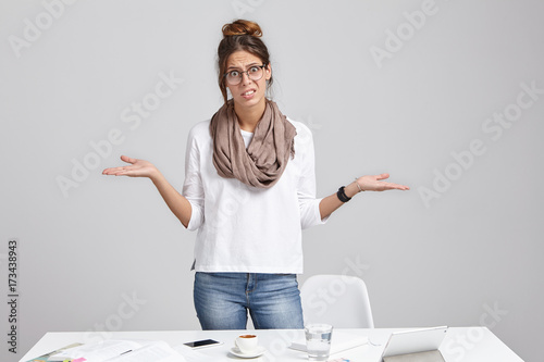 Unhappy woman frowns face in displaesure, shrug shoulders, has bad mood, stands at office table, makes mistake in annual report, should do calculation from beginning, look desperatedly and upset