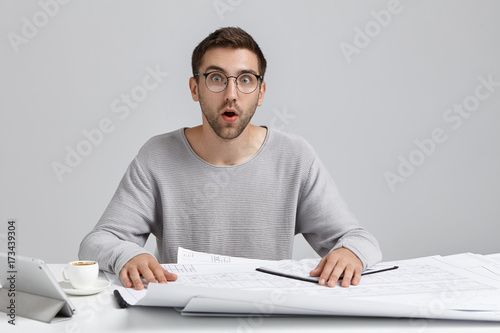 Puzzled surprised young office worker realizes his fault, doesn`t want to create construction plan from beginning, finds out that he should present it today, has no time and strength for improvement photo