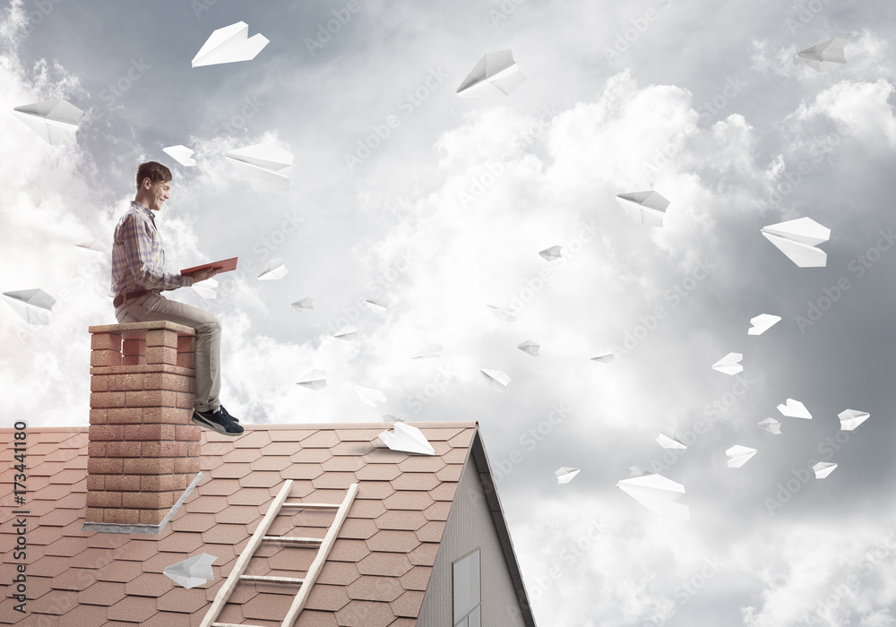 Man on brick roof reading book and paper planes flying in air Man on brick roof reading book and paper planes flying in air