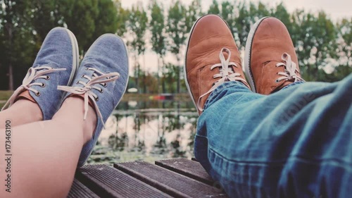 Point Of View: Couple sitting on a wooden jetty playing footsie, close up on modern hipster shoes. 4K Ultra HD. Relaxed time by the lake on a pier. POV: romantic young love by the lakeside. Fashion.  photo