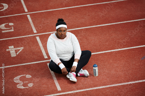 Cross-legged overweight woman in activewear having break for drink after workout on stadium