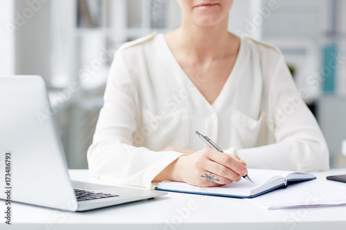 Unrecognizable young businesswoman wearing white shirt sitting at office desk and taking necessary notes, blurred background © pressmaster
