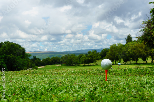 close up of white golf ball on orange tee on green grass with blue sky and cloud. copy space for your text.