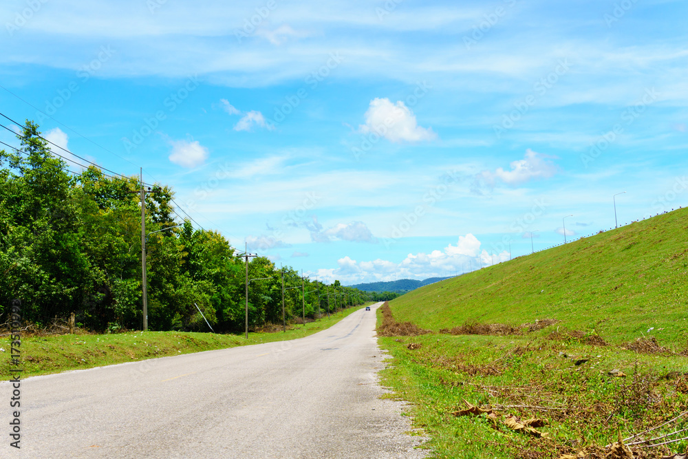 empty asphalt country road along the wall of dam with green grass and blue sky with clouds and mountain background in countryside