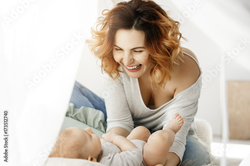 Portrait of young attractive mother laughs and playing with newborn child in comfy light bedroom. Warm mornings with family. Happy childhood.
