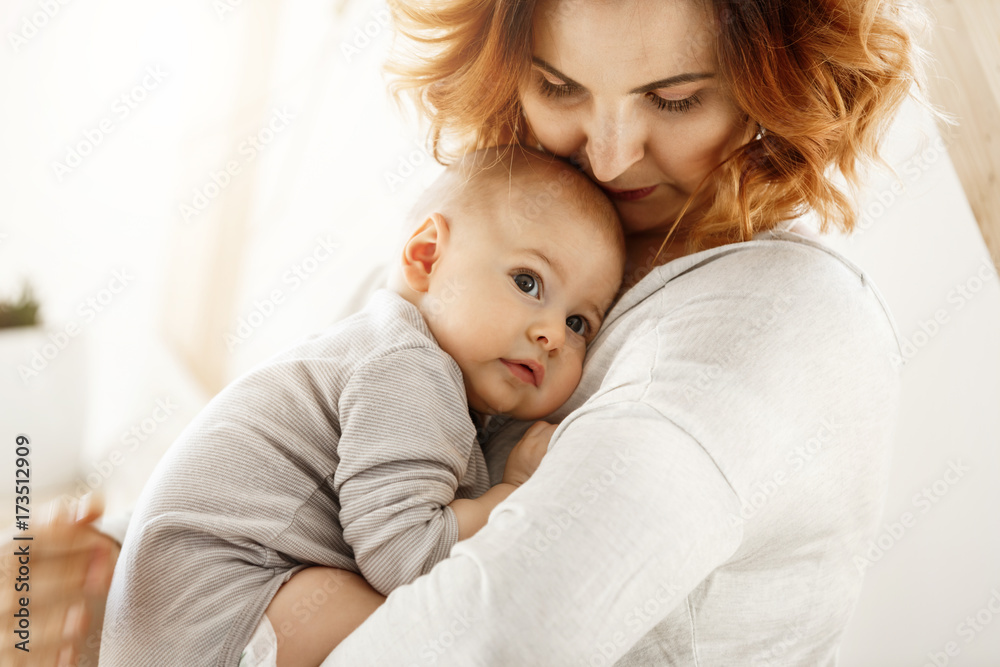 Beautiful young mother tenderly hugs her little cute child. Mom looking at her kid and smiling while baby looks aside and snuggling to mom chest. Happy family moments. Beautiful young mother tenderly hugs her little cute child. Mom looking at her kid and smiling while baby looks aside and snuggling to mom chest. Happy family moments.