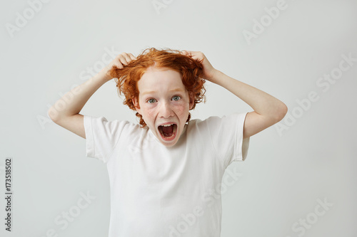 Foto Headshot of cute surprised little boy with freckles looking in camera with mad expression and opened mouth, holding hair with hands
