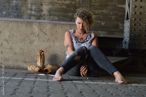 a hopeless punk woman under a bridge with bottles of alcohol and other drugs around her 