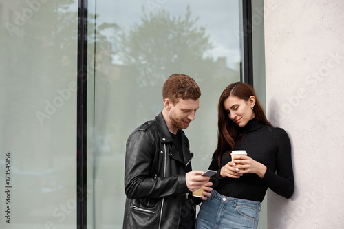 Isolated outdoor picture of modern cool young couple talking outside building with large window: brunette woman drinking takeawy coffee while bearded guy showing her something on mobile phone photo