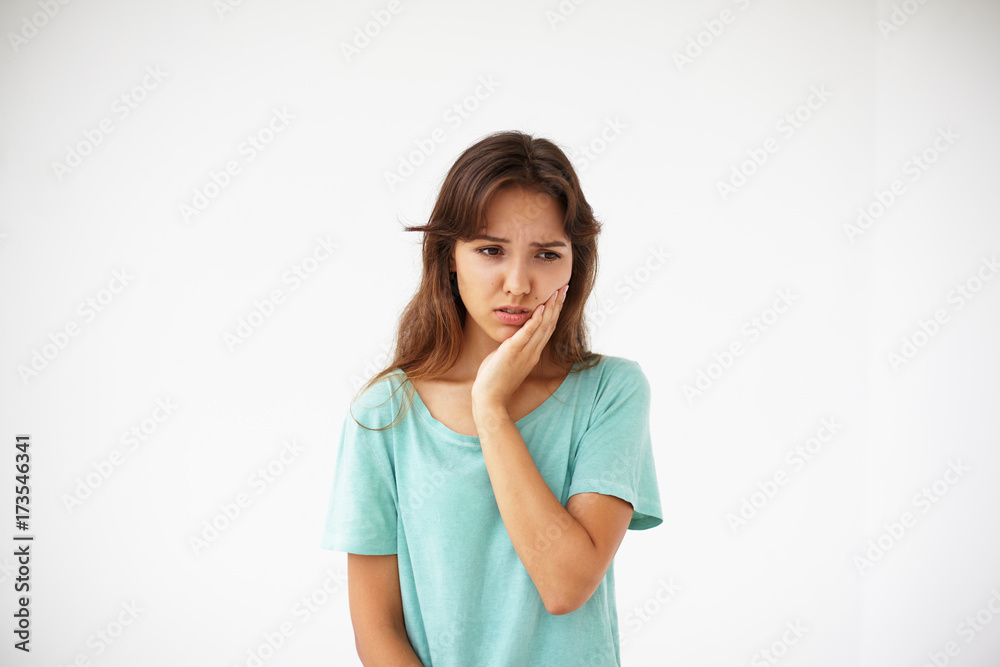 Unhappy cute young brunnete female with long dark hair holding hand on her cheek while suffering from toothache. Pretty girl touching face having painful look after she had her wisdom tooth out Unhappy cute young brunnete female with long dark hair holding hand on her cheek while suffering from toothache. Pretty girl touching face having painful look after she had her wisdom tooth out